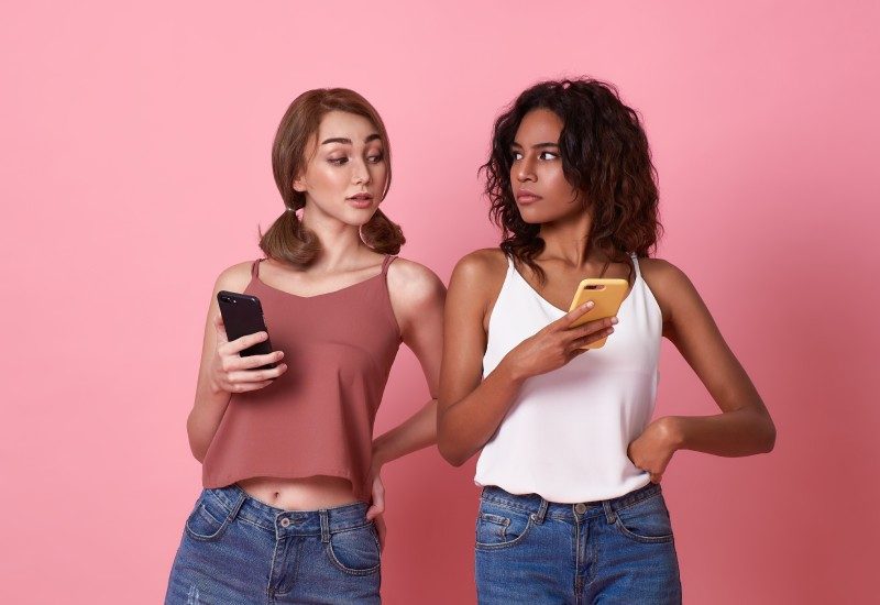 Reasons To Keep Your Social Media Profile Private 1 Two young women stand next to each other holding smartphones. One of them peeks over the other's shoulder to read her screen.